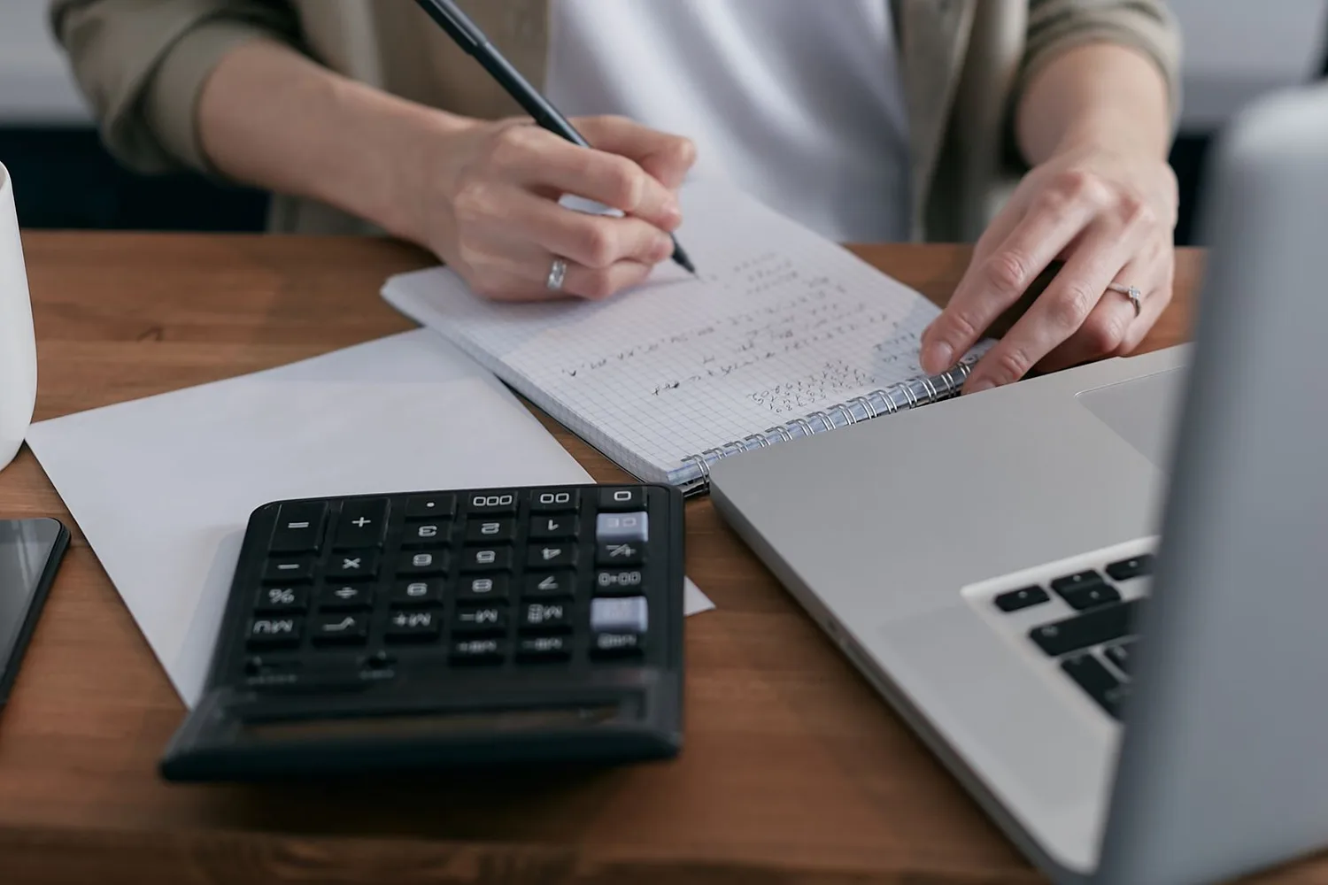 A small-business owner working out costs by hand with a calculator, a laptop, and a notebook of figures on a wooden desk.